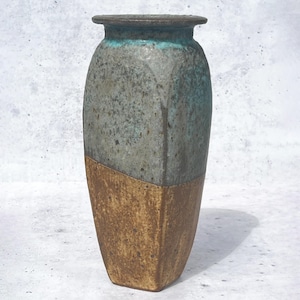 May include: A tall, square-shaped ceramic vase featuring a two-tone glaze. The upper portion displays a mottled grey and teal finish, while the lower section is a textured, earthy brown. The vase has a wide, flat rim and a sturdy, angular base.