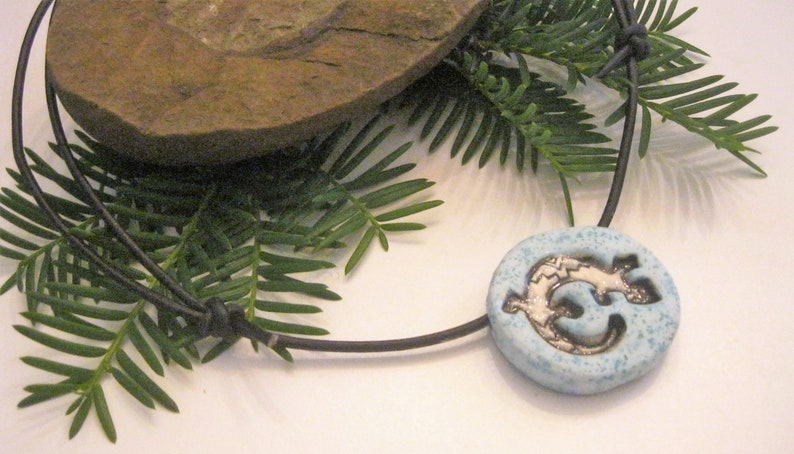 Puede incluir: Un collar de cuero negro con un colgante de cer&aacute;mica azul con un dise&ntilde;o plateado de un p&aacute;jaro estilizado.