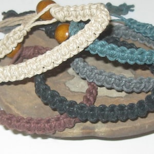 May include: Five braided hemp bracelets in brown, black, gray, teal, and natural colors. The bracelets are stacked on top of each other and have wooden beads.