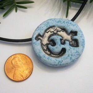 Puede incluir: Un collar con un colgante de cer&aacute;mica azul con un dise&ntilde;o de lagarto blanco. El colgante es aproximadamente del tama&ntilde;o de una moneda de un centavo de euro.