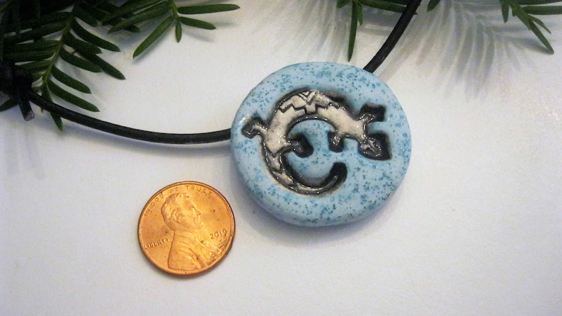 Puede incluir: Un collar con un colgante de cer&aacute;mica azul con un dise&ntilde;o de lagarto blanco. El colgante es aproximadamente del tama&ntilde;o de una moneda de un centavo de euro.