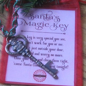 May include: A silver-colored key with a Santa Claus design, attached to a red and green braided cord, rests on a pink card inside a red mesh bag. The card reads "Santa's Magic Key."
