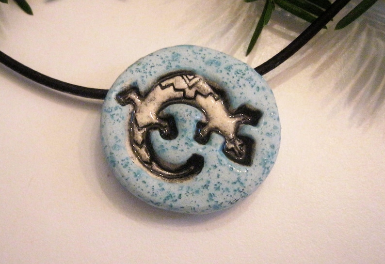 Puede incluir: Un collar con un colgante de cer&aacute;mica azul con un dise&ntilde;o de lagarto blanco. El lagarto est&aacute; enroscado en un c&iacute;rculo y tiene detalles negros.