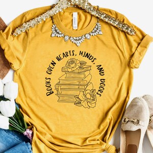 May include: A mustard yellow t-shirt with a black graphic design of a stack of books with roses and the text "Books open hearts, minds, and doors".
