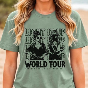 May include: Sage green t-shirt with the text "LIGHT IT UP WORLD TOUR" and illustrations of two women wearing sunglasses. The phrase "THROUGH LOVE, ALL IS POSSIBLE" is printed below the main text.
