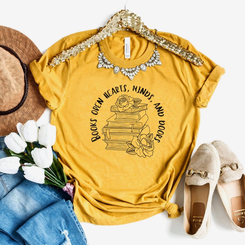 May include: A mustard yellow t-shirt with a black graphic design of a stack of books with roses and the text "Books open hearts, minds, and doors".