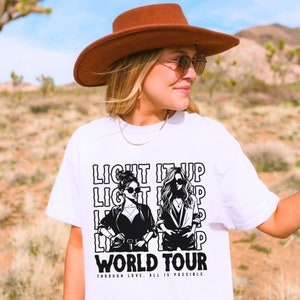 May include: White t-shirt with a black and white graphic of two women wearing sunglasses. The text "LIGHT IT UP WORLD TOUR" is printed above the graphic, with the phrase "THROUGH LOVE, ALL IS POSSIBLE" below.