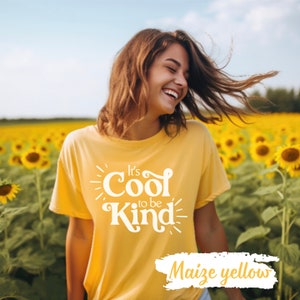 Pode incluir: Uma mulher vestindo uma camiseta amarela com o texto "It's Cool to be Kind" em branco. A camiseta está em um campo de girassóis. O texto "Maize yellow" está escrito em branco sobre um fundo amarelo.
