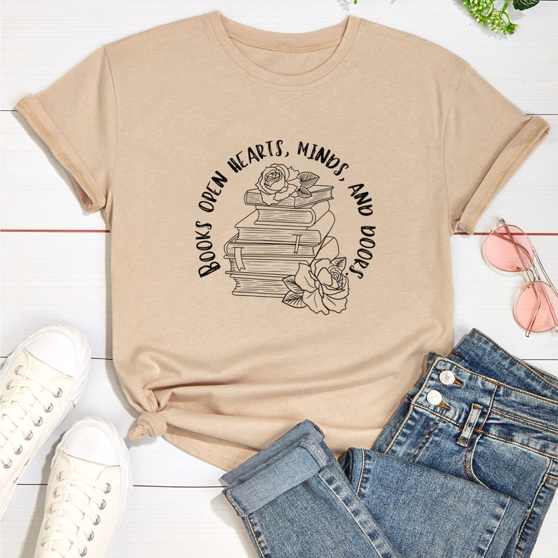 May include: A beige t-shirt with a graphic of a stack of books and roses with the text "Books open hearts, minds, and doors".