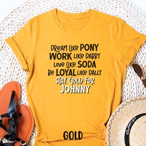 K&ouml;nnte beinhalten: Gelbes T-Shirt mit schwarzem Text, der "Dream like Pony, Work like Darry, Love like Soda, Be Loyal like Dally, Stay Gold for Johnny" lautet. Das Wort "Gold" ist in Schwarz am unteren Rand des Shirts gedruckt.