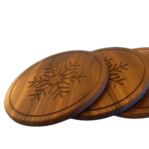 May include: Four oval-shaped wooden coasters with a snowflake design carved into the surface. The coasters are made of a dark brown wood and have a groove around the edge. The coasters are arranged in a row, slightly overlapping.