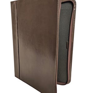 May include: A brown leather portfolio with a zip closure. The portfolio is open, revealing a black interior. The exterior has a vertical seam down the front. The zip is brown.