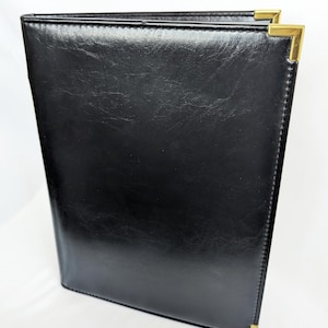 May include: A black menu cover or binder with a smooth, slightly glossy finish and stitched edges. The rectangular item features polished gold-tone metal corner protectors on the top and bottom right corners, providing a structured appearance.