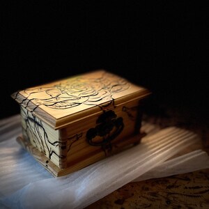 May include: A wooden box with a black rose design on the lid. The box has a metal handle and is sitting on a white surface.