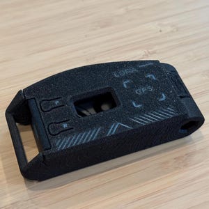 May include: A black rectangular device with the text "LORA" and "GPS" printed on the top. The device has a textured surface and a small rectangular opening. The device is on a light brown wooden surface.