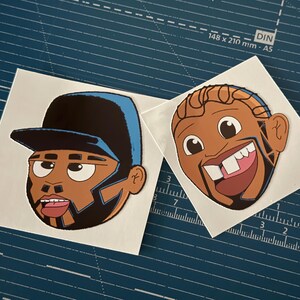 May include: Two cartoon stickers featuring Black men's faces. One wears a black cap with blue accents, the other has a tooth missing. Both have brown skin, black beards, and stylized features. The stickers are on a blue cutting mat.