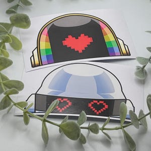 May include: Two stickers featuring a helmet design. One has a black face with a pixelated red heart and rainbow accents. The other has a blue face with pixelated red heart-shaped eyes.