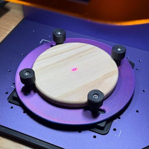 May include: A close-up of a laser engraving machine with a wooden disc in the center. The machine has a purple base and a circular purple platform. Four black knobs secure the wooden disc. A red laser dot is visible on the wood.