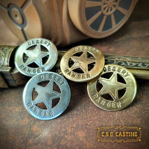 May include: Four metal Desert Ranger badges in silver and gold tones. Each badge features a star in the center and the words "Desert Ranger" around the edge. The badges are displayed on a brown surface with a blurred background.