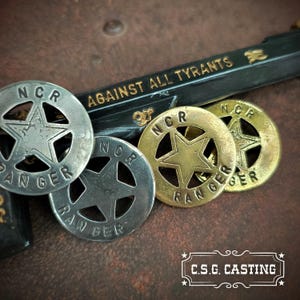 May include: Four metal NCR Ranger badges in silver and gold tones are displayed. Each badge has a star design and the words "NCR RANGER". A gun with the words "AGAINST ALL TYRANTS" is in the background. The image also includes the text "C.S.G. CASTING".