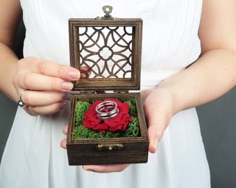 Puede incluir: Una caja de madera para anillos con un diseño tallado en la tapa. La caja está abierta y contiene una rosa roja con dos anillos plateados en su interior. La caja está siendo sostenida por una persona.