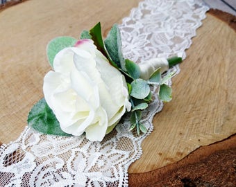 May include: A white faux rose with green leaves and a white lace ribbon on a wooden slice.