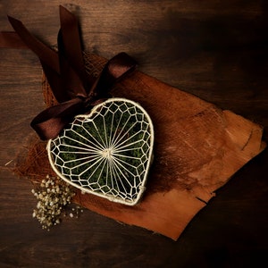 May include: A white heart-shaped wire frame filled with green moss and tied with a brown ribbon on a brown wooden surface.