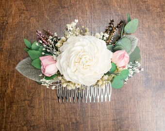 May include: A floral hair comb featuring a large, cream-colored flower as the focal point, surrounded by pink and green accents. The comb includes a variety of dried flowers and greenery, with a silver comb base. The arrangement is set against a dark wood surface.
