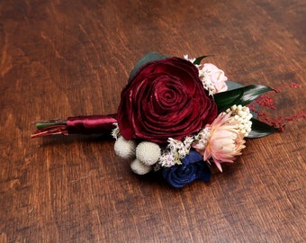 May include: A burgundy and navy blue flower bouquet with white and pink accents. The bouquet is made of silk flowers and is tied with a burgundy ribbon.
