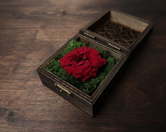 Puede incluir: Una caja de madera con una rosa roja y musgo verde en su interior. La caja tiene un diseño decorativo e intrincado en la tapa.