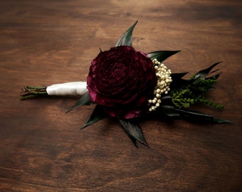 May include: A burgundy ranunculus flower bouquet with white berries and green foliage, tied with a white ribbon.