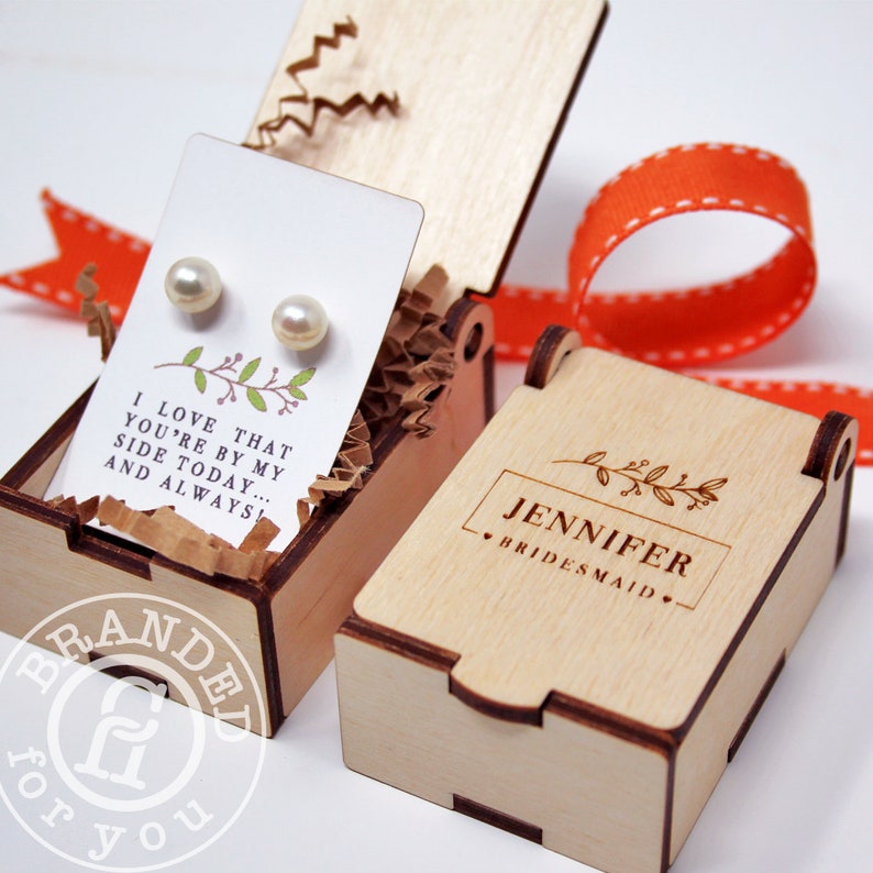 May include: A wooden gift box with a hinged lid, containing a pair of pearl earrings on a card with text. The box is engraved with the name "Jennifer" and the word "Bridesmaid". An orange ribbon is in the background.