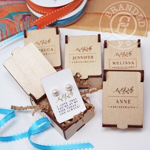 May include: Wooden gift boxes with engraved names and floral designs. One box is open, revealing a card with pearl earrings and the text "I LOVE THAT YOU'RE BY MY SIDE TODAY... AND ALWAYS!". Ribbons and decorative paper are in the background.