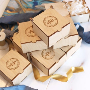May include: A stack of six small, wooden boxes with engraved initials. The boxes are light brown and have a natural wood finish. The initials are in a circle with a decorative border.