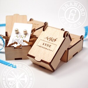 May include: Wooden gift boxes with engraved details. One box is open, revealing pearl earrings on a card with text. The box lids are engraved with a name and the word "Bridesmaid". The boxes are filled with crinkle paper.