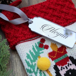 May include: A red and white knitted Christmas stocking with a nutcracker design. A silver tag with the name "Aleyna" is attached to the stocking.