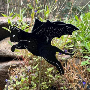 May include: A black metal cat garden stake with bat wings, outlined in white, is displayed in a flower pot. The cat is in mid-flight, with its wings spread. The background features green plants and a stone.