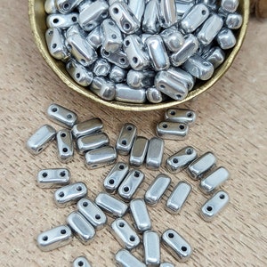 Könnte beinhalten: Eine Sammlung glänzender, silberfarbener Metallperlen. Die Perlen sind rechteckig mit abgerundeten Kanten und zwei Löchern zum Auffädeln. Einige Perlen befinden sich in einer kleinen, goldfarbenen Schale, andere sind auf einer Holzoberfläche verstreut. Ideal für die Schmuckherstellung.