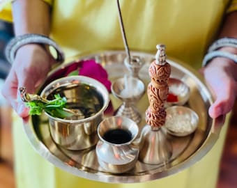 May include: A silver tray containing a selection of items, including a small bowl with green leaves, a small bowl with a dark liquid, a small bowl with white powder, a silver incense holder, and a wooden bell with silver accents.