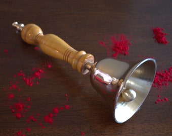 May include: A silver hand bell with a wooden handle. The bell is lying on a dark brown surface with red powder scattered around it.