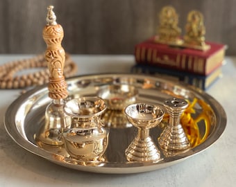 May include: A silver tray with a wooden handled bell, a small silver bowl, a silver diya, and a silver incense burner. The tray is on a light grey surface.
