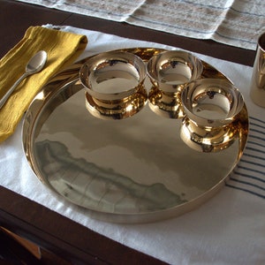 May include: A gold-toned serving tray with three smaller bowls, a glass, and a yellow napkin with a silver spoon. The tray and bowls are reflective and have a shiny finish.
