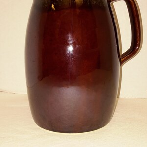 May include: A brown ceramic pitcher with a glazed finish and a light brown and yellow rim. The pitcher has a rounded body and a handle.
