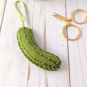 Puede incluir: Adorno de pepinillo de fieltro verde hecho a mano con detalles bordados y un lazo de cinta verde. Tijeras doradas y un tarro de Kosher Dill Pickle Spears en el fondo. El adorno mide aproximadamente 10 cm de largo.