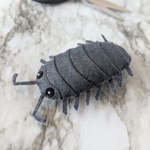 Op de afbeelding: Een handgemaakt grijs vilten pissebed-speeltje met zwarte ogen en antennes. Het gesegmenteerde lichaam is gedetailleerd met gestikte lijnen. Een open schaar staat op de achtergrond, wat wijst op een knutselproject.