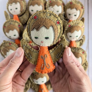 Puede incluir: Un grupo de adornos de ángeles de fieltro con cabello castaño, rostros blancos y vestidos naranjas. Los ángeles tienen alas doradas y están decorados con cuentas rojas. Los adornos están hechos a mano y serían una hermosa adición a cualquier árbol de Navidad.