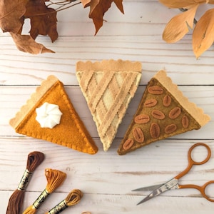 Könnte beinhalten: Drei dreieckige Tortenstücke aus Filz in Orange, Creme und Braun. Das orangefarbene Stück hat einen weißen Filzklecks, das cremefarbene Stück ein Gittermuster und das braune Stück ovale Filzakzente. Enthält Stickgarn und eine Metallschere.