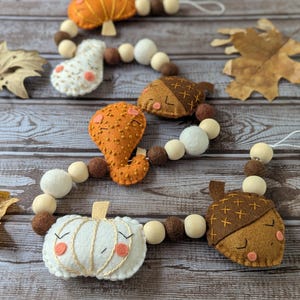 May include: A fall-themed garland featuring felt pumpkins, acorns, and other autumn shapes. The garland includes white, orange, and brown felt pieces, with wooden beads. The pumpkins and acorns have stitched details and smiling faces.