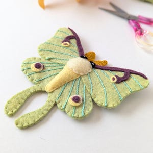May include: A handmade felt moth with a light green body, purple antennae, and yellow accents. The moth has detailed stitching and a whimsical design. The wings are a light green color with blue stitching. The moth is a unique craft item.
