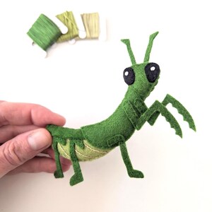 May include: A handmade green felt praying mantis with large black eyes and detailed legs. The insect is held by a hand, showcasing its intricate design. The background includes green thread spools and greenery, suggesting a craft project.